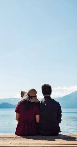 couple sitting on dock, facing body of water and mountains